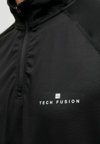 Must musta, millel on sile ja veniv kangas. Omab kõrget kraed ja rinnal on valge "TECH FUSION" logo.