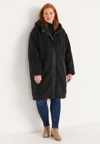 Zwarte, zachte, oversized jas met een capuchon en drukknoopsluiting, met zijzakken en een zachte textuur, gedragen over donkerblauwe spijkerbroek.