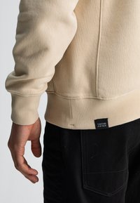 Beige fleece sweatshirt met ribgebreide manchetten, met een zijnaaddetail en een klein zwart merklabel aan de onderrand. Zwarte broek zichtbaar.
