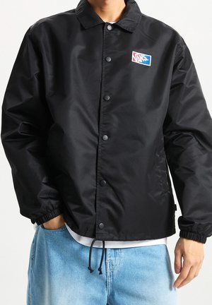 Personne portant une veste noire Vans avec des boutons-pression et des poignets élastiques, associée à un jean bleu clair et une chemise blanche qui dépasse sous la veste.