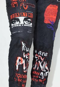 Schwarze Stoffhosen mit einer zerknitterten Textur, verziert mit auffälligem roten und weißen Grafiktext und Bildern, einschließlich Gesichtern und Designelementen.