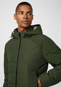 Chaqueta acolchada verde con capucha, que presenta un diseño estructurado, cremallera negra, bolsillos laterales y un forro beige. Tejido texturizado con un patrón sutil.