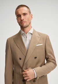Beige Doppelreiher-Blazer mit strukturierter Oberfläche, Reverskragen und braunen Knöpfen, kombiniert mit einem hellgrauen Hemd.