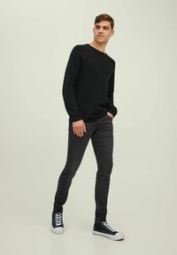 Svart genser med rund hals, grå smale jeans og svarte høyhalse sneakers. Modellen står mot en lys grønn bakgrunn.