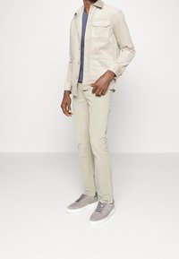 Beżowa kurtka z korduroju z jedną kieszenią na piersi, noszona na granatowej koszulce, w komplecie z jasnobeżowymi spodniami typu slim fit i szarymi zamszowymi sneakerami.