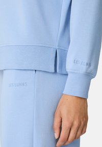 Suéter azul claro con puños y dobladillo acanalados, que presenta un logo bordado "LES LUNES" en la manga y un detalle de abertura lateral en el dobladillo.