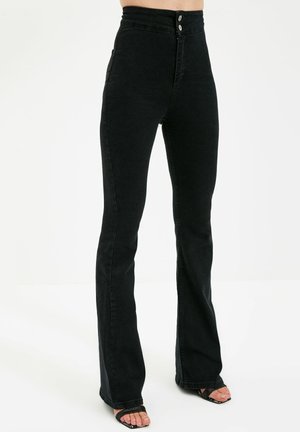 Czarne, wysokiego stanu, dopasowane dżinsy z lekkim rozszerzeniem, noszone z czarnymi sandałami na odkryte palce, pokazane od talii do stóp na białym tle.