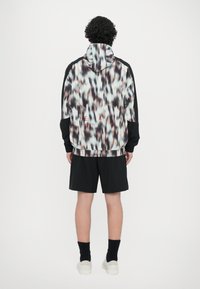 Jachetă ușoară multicoloră cu guler înalt și fermoar, având un model de linii estompate, purtată cu pantaloni scurți negri și adidași albi.