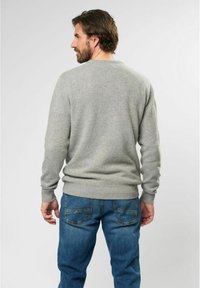 Hombre con barba que lleva un suéter gris y jeans azules, de pie de espaldas contra un fondo claro y liso.