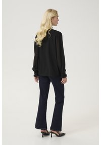 Črna prozorna bluzka s napihnjenimi rokavi, v kombinaciji z temno modrimi širokimi hlačami in črnimi visokimi sandalami. Glasovna tekstura, minimalističen dizajn.