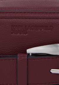 Burgundijas ādas josta ar iespiesto "KARL LAGERFELD JEANS" logo. Iezīmes sudraba metāla sprādze un kontrastējošas melnas šuves.