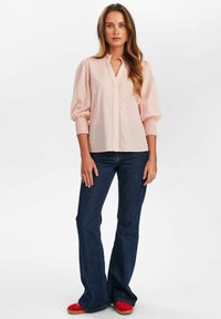 Blusa rosa chiaro con maniche a palloncino e scollatura a V, caratterizzata da un piccolo motivo geometrico, abbinata a jeans svasati blu scuro e scarpe rosse.