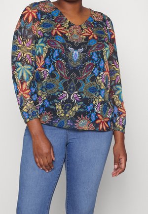 Femme portant un chemisier à manches longues et col en V avec des motifs floraux et cachemire éclatants, assorti à un jean bleu.