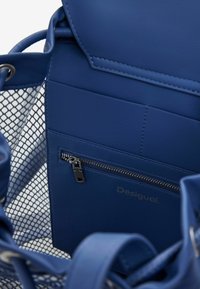 Blaues Kunstledertasche mit Netzdetails, ausgestattet mit einer vorderen Reißverschlusstasche und geprägtem Logo. Glatte Textur und strukturiertes Design.