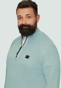 Jasnoniebieski, teksturowany sweter zapinany na zamek z wysokim kołnierzem, z czarną naszywką logo i kontrastującym detalem zamka.