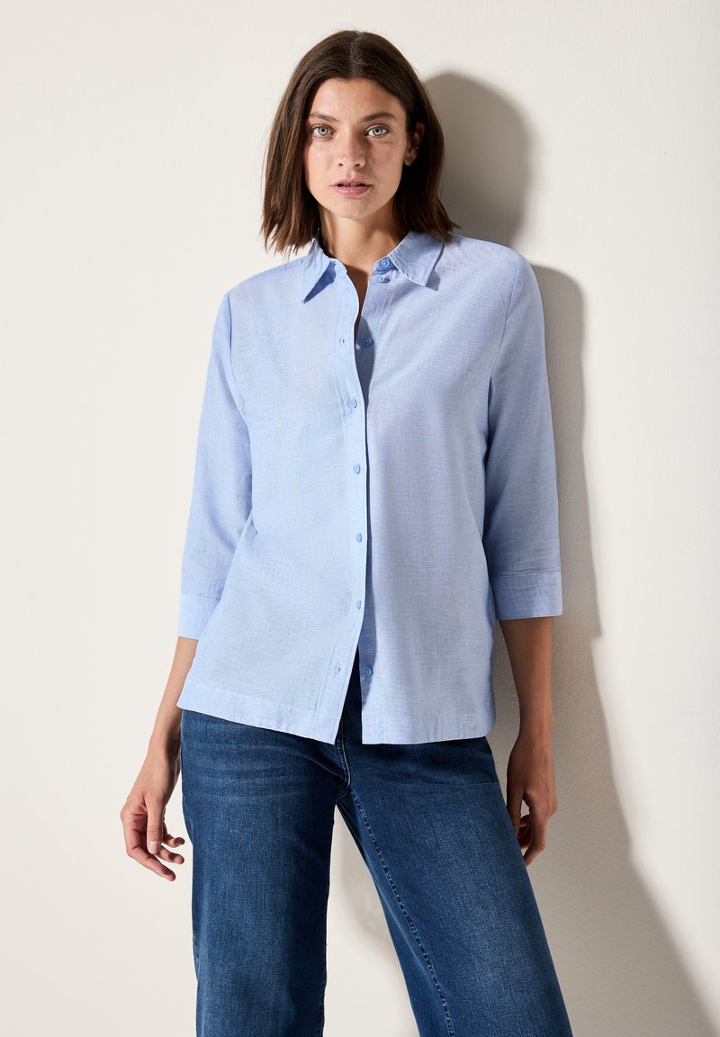 Camicia button-up azzurro chiaro con colletto, maniche a tre quarti e una consistenza morbida, abbinata a jeans blu scuro.