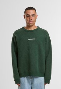 Zeleni pleteni pulover s teksturiranim završetkom, s okruglim izrezom i spuštenim ramenima. U središtu se nalazi bijeli natpis "ANOTHER COTTON."