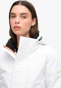 Witte waterdichte jas met zwarte voering, oranje rits, reliëf logo-detail op de voorkant en verstelbare capuchon met hardware accenten.