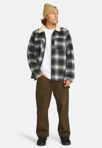 Man staat in een geruite flanellen shirt met een bontkraag, beige beanie, witte t-shirt en bruine broek, tegen een effen achtergrond.