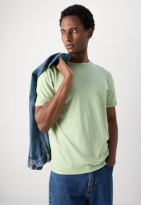 Tricou din bumbac verde deschis, cu guler rotund și mâneci scurte, purtat cu o jachetă din denim aruncată pe umăr. Se poartă blugi albaștri.