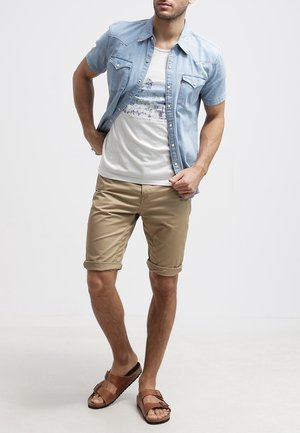 Hellblaues Jeanshemd mit kurzen Ärmeln, weißes T-Shirt mit Grafikdruck, beige Shorts und braune Sandalen, mit hochgekrempelten Ärmeln und lockerem Schnitt.