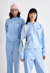 Dos modelos vistiendo sudaderas y pantalones ligeros azules a juego; uno con un gorro rosa y el otro llevando una bolsa de mano crema sobre un fondo blanco.