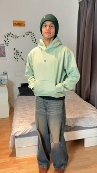 Young man wearing a green beanie and hoodie stands with hands in pockets in a bedroom with a bed, curtains, and a Los Angeles sign on the wall.