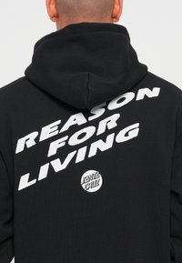 Črna kapuca z izrazitim belim napisom "RAZLOG ZA ŽIVLJENJE" in okroglo "SANTA CRUZ" logotipom na hrbtni strani. Mehka tkanina in zasnova s kapuco.