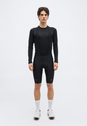 Mladić stoji, obučene crne biciklističke bretelice s dugim rukavima, bijele čarape i bijele biciklističke cipele, na jednostavnoj bijeloj pozadini.