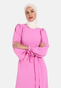 Lyserød kjole med pufærmer, indsnørede ærmer og taljebånd. Stoffet ser blødt og struktureret ud. Kappen er ledsaget af en flødefarvet hijab.