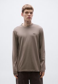 Hosszú ujjú barna pamut póló kerek nyakkivágással és egy kis fekete logóval a mellkason. Sima textúrájú, kényelmes szabású.