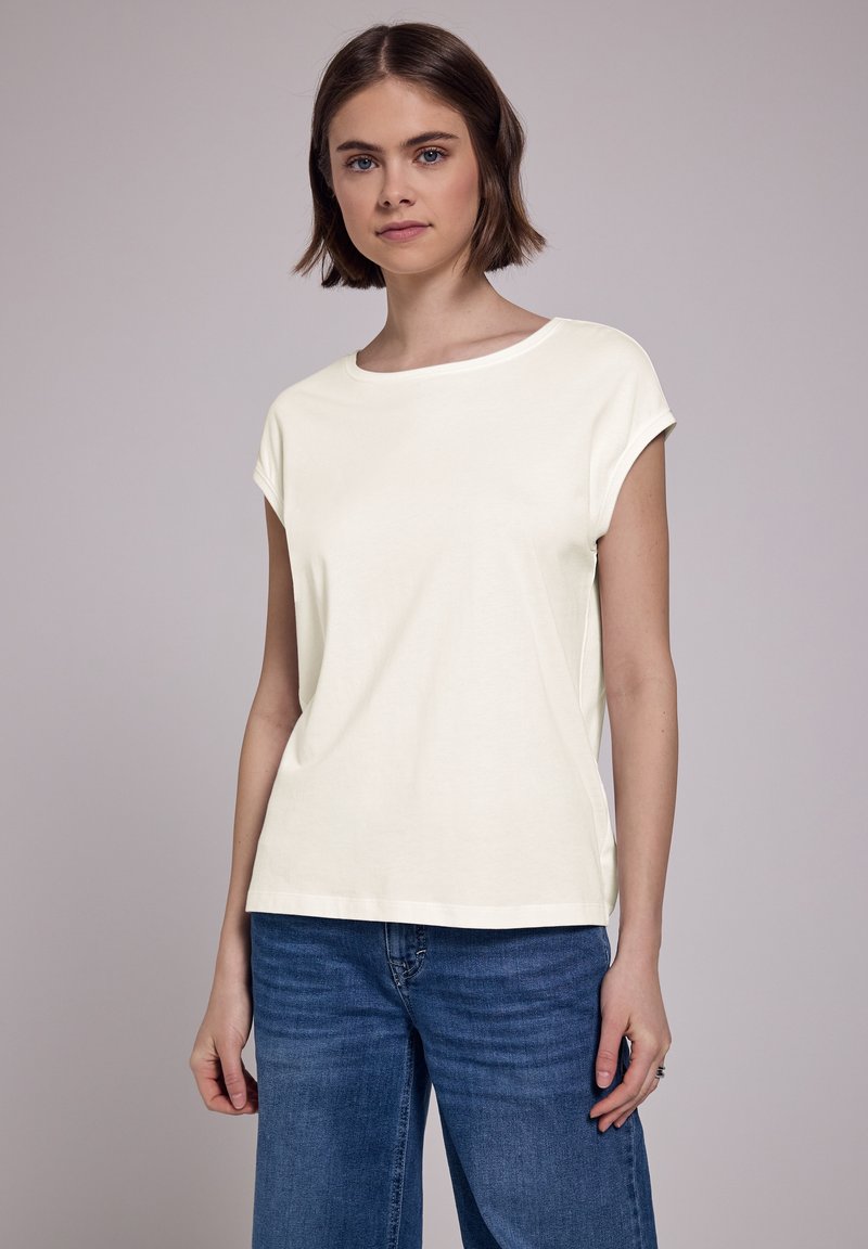 Cremefarvet bomulds-T-shirt med korte ærmer, rund halsudskæring og en løs pasform. Kombineret med blå straight-leg jeans.