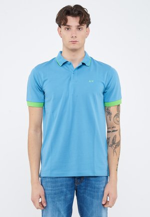 Jonge man staat frontaal, draagt een blauw poloshirt met groene bies en blauwe spijkerbroek, toont tatoeages op zijn rechteronderarm.