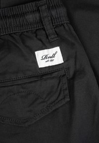Black fabric pants with elastic waistband, back pocket, and white rectangular label reading "Reell est. 1997" stitched above pocket.