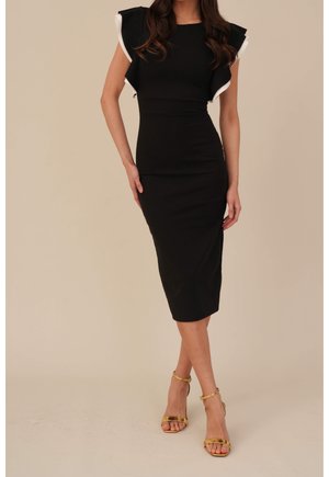 Femme en robe midi noire avec des manches à volants bordées de blanc et des talons hauts dorés à bride à la cheville, debout jambes croisées.