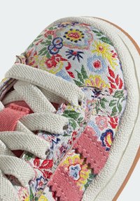 Gros plan sur l'avant de la sneaker avec des lacets blancs, un tissu floral multicolore et une rayure en daim rose en zigzag le long de la semelle texturée.