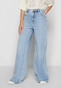 Personne portant un jean large bleu clair et des baskets blanches, debout avec une main dans la poche et des tatouages visibles sur l'avant-bras.