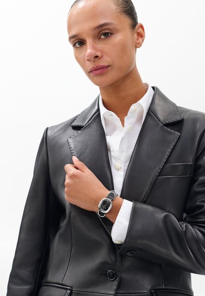 Schwarzer Lederblazer mit ausgeprägten Revers, getragen über einem weißen Hemd mit Knöpfen; eine silberne Uhr mit schwarzem Zifferblatt ergänzt das Outfit.