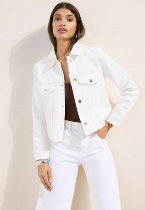 Junge Frau, die eine weiße Jacke mit Knöpfen, weiße Hosen und ein braunes Oberteil trägt, steht mit einer Hand in der Tasche vor einem einfarbigen Hintergrund.