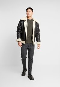 Schwarze Leder-Shearling-Jacke mit cremefarbener Kragen und Bündchen, kombiniert mit einem grünen strukturierten Pullover und grauen Karohosen. Schwarze Schuhe vervollständigen den Look.