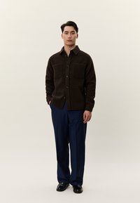 Vyras stovi vilkėdamas tamsiai rudą marškinių švarką su saga, tamsiai mėlynas kelnes ir juodus elegantiškus batus priešais paprastą šviesų foną.