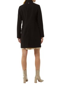 Schwarzer Wollmantel mit hohem Kragen, Ein-Knopf-Verschluss und schlankem Rückendesign. Getragen über einem hellen Kleid und beigen Knöchelschuhen.