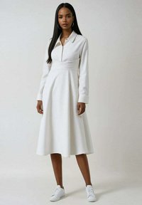 Femme portant une robe midi blanche à manches longues et col, avec une ceinture à la taille ajustée, assortie à des baskets blanches, se tenant devant un fond uni.