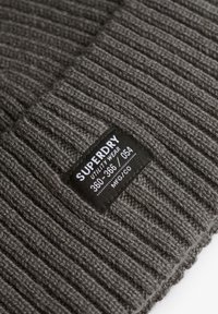 Bonnet en maille côtelée gris charbon avec une étiquette noire indiquant "SUPERDRY" et des détails d'entretien, présentant un tissu texturé et des coutures visibles.