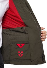 Oliwkowa kurtka z czerwonym siateczkowym podkładem, bocznym zamkiem błyskawicznym i kieszenią na piersi z kontrastującymi czerwonymi akcentami oraz haftem.