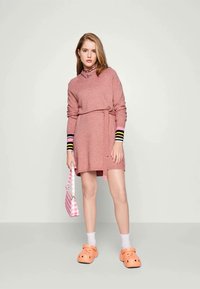 Ροζ πλεκτό φόρεμα με μακριά μανίκια και ζώνη, που διαθέτει πολύχρωμες ριγέ μανσέτες. Συνδυασμένο με πορτοκαλί κλόουν και ροζ καρό τσάντα.