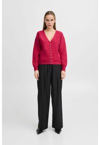 Gaiši rozā kardigāns ar V veida kakla izgriezumu, pogu aizdari un divām priekšējām kabatām. Saskaņots ar melniem platām biksēm un melniem apaviem.