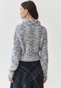 Knit sweater in blue and white with cowl neck. Textured fabric with long sleeves. Paired with a navy plaid skirt featuring colorful lines.