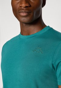Teal lühikeste varrukatega T-särk, mis on valmistatud pehmest kangast, ümarate kaelusteni ja väikese tikandiga logoga vasakul ülal rinnal.