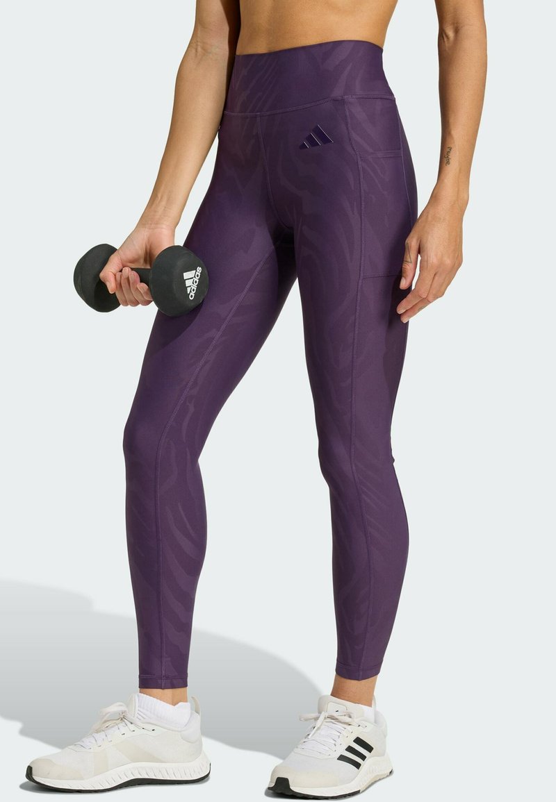 Lila Leggings mit hoher Taille, die ein tonales Muster und eine enganliegende Passform aufweisen, gefertigt aus einem dehnbaren, atmungsaktiven Material. Hantel wird in einer Hand gehalten.